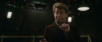 Movie still from “Luther: The Fallen Sun” (2023), directed by Jamie Payne – An older man wearing a brown jacket holding a microphone; Close Up shot, Over the shoulder angle