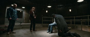 Movie still from “Luther: The Fallen Sun” (2023), directed by Jamie Payne – Two men sit on a chair and one man is kneeling on the ground; Wide shot, Low angle