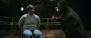 Movie still from “Luther: The Fallen Sun” (2023), directed by Jamie Payne – A man holding an object in front of a woman sitting in a chair; Medium shot, Over the shoulder angle