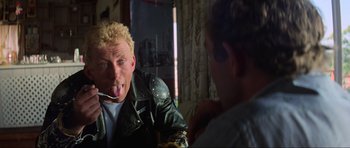 Movie still from “Mad Max” (1979), directed by George Miller – A man with blonde spiky hair is eating something off a spoon; Close Up shot, Over the shoulder angle