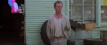 Movie still from “Mad Max” (1979), directed by George Miller – A man standing in front of a wall with a tire; Medium shot, Low angle