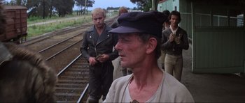 Movie still from “Mad Max” (1979), directed by George Miller – A group of men standing next to each other on train tracks; Medium shot, Over the shoulder angle