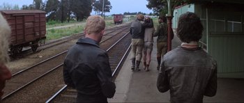 Movie still from “Mad Max” (1979), directed by George Miller – A group of people walking along a train track; Wide shot, Over the shoulder angle