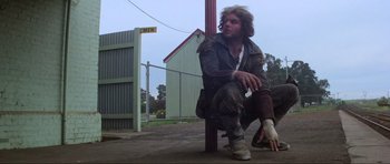 Movie still from “Mad Max” (1979), directed by George Miller – A man sitting on the ground in a parking lot; Wide shot, Low angle