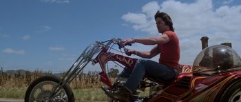 Movie still from “Mad Max” (1979), directed by George Miller – A man riding a motorcycle on the side of the road; Medium shot, Low angle