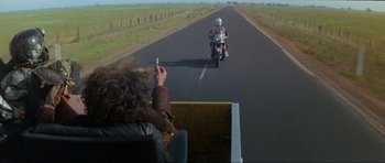 Movie still from “Mad Max” (1979), directed by George Miller – A person is taking a picture of a person riding a motorcycle on the road; Wide shot, High angle