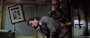 Movie still from “Mad Max” (1979), directed by George Miller – A man in a suit is being held by another man in a leather jacket; Medium shot, Over the shoulder angle
