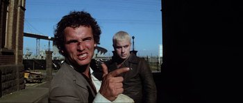 Movie still from “Mad Max” (1979), directed by George Miller – Two men are posing for a picture while one of them is pointing at the camera; Medium shot, Low angle
