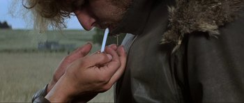 Movie still from “Mad Max” (1979), directed by George Miller – A man is smoking a cigarette while a woman is putting something in his mouth; Close Up shot, Over the shoulder angle