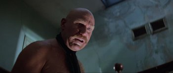 Movie still from “Mad Max” (1979), directed by George Miller – A man with a shaved head and a black tie; Close Up shot, Low angle