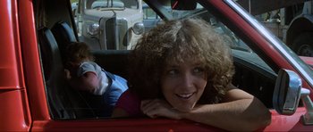 Movie still from “Mad Max” (1979), directed by George Miller – A woman with curly brown hair sitting in a red car; Close Up shot, Over the shoulder angle