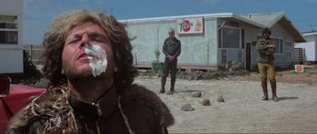 Movie still from “Mad Max” (1979), directed by George Miller – A man is eating a piece of cake while another man watches; Medium shot, Over the shoulder angle