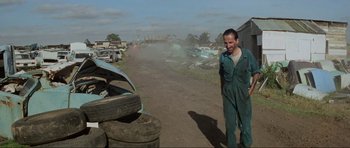 Movie still from “Mad Max” (1979), directed by George Miller – A man standing next to a pile of junk on the side of the road; Wide shot, High angle