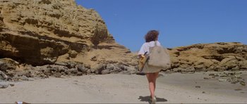 Movie still from “Mad Max” (1979), directed by George Miller – A woman walking on the beach carrying a bag; Wide shot, Low angle