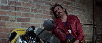 Movie still from “Mad Max” (1979), directed by George Miller – A man leaning against a brick wall while holding a motorcycle; Close Up shot, Low angle