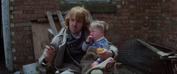 Movie still from “Mad Max” (1979), directed by George Miller – A man and a child sitting next to a brick wall; Medium shot, Low angle