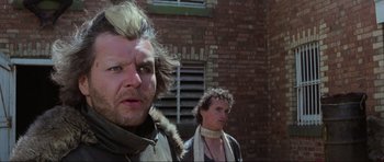 Movie still from “Mad Max” (1979), directed by George Miller – Two men standing next to each other in front of a brick building; Close Up shot, Low angle