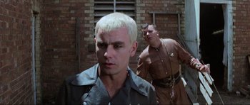 Movie still from “Mad Max” (1979), directed by George Miller – A man with white hair is standing next to another man; Close Up shot, Low angle