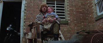 Movie still from “Mad Max” (1979), directed by George Miller – A man holding a baby while sitting on a chair; Medium shot, Low angle