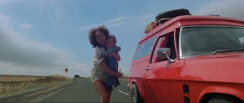 Movie still from “Mad Max” (1979), directed by George Miller – A woman holding a child next to a red van; Wide shot, Low angle