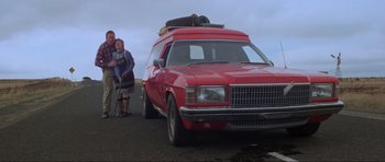 Movie still from “Mad Max” (1979), directed by George Miller – Two people standing in front of a red car; Wide shot, Low angle