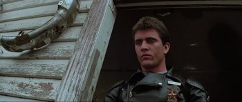 Movie still from “Mad Max” (1979), directed by George Miller – A man in a leather jacket standing next to a wooden post; Close Up shot, Low angle