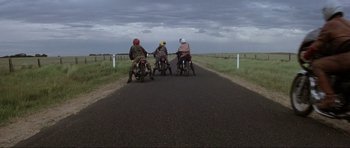 Movie still from “Mad Max” (1979), directed by George Miller – A group of people riding motorcycles down the middle of a road; Extreme Wide shot, Low angle