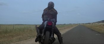Movie still from “Mad Max” (1979), directed by George Miller – A man riding a motorcycle down the middle of a road; Wide shot, Low angle