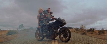 Movie still from “The Road Warrior” (1981), directed by George Miller – A couple of people on a motorcycle on the side of the road; Wide shot, Low angle