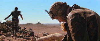Movie still from “The Road Warrior” (1981), directed by George Miller – A man in a helmet and goggles looking down at a rock formation; Wide shot, Over the shoulder angle