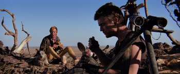 Movie still from “The Road Warrior” (1981), directed by George Miller – Two men sitting on the ground and one of them is holding a camera; Medium shot, Low angle