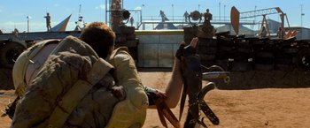 Movie still from “The Road Warrior” (1981), directed by George Miller – A person riding a bike on top of a dirt field; Wide shot, Over the shoulder angle