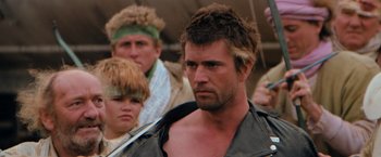 Movie still from “The Road Warrior” (1981), directed by George Miller – A group of men standing next to each other; Close Up shot, Over the shoulder angle