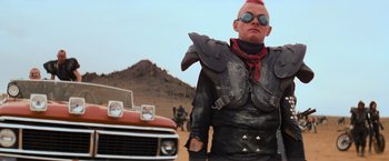 Movie still from “The Road Warrior” (1981), directed by George Miller – A man in a leather jacket standing in front of a red truck; Medium shot, Low angle