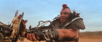 Movie still from “The Road Warrior” (1981), directed by George Miller – A man with a mohawk is holding a crossbow in his hand; Close Up shot, Low angle
