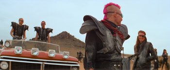 Movie still from “The Road Warrior” (1981), directed by George Miller – A man with a pink mohawk stands in front of a red car; Medium shot, Over the shoulder angle