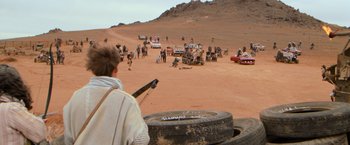 Movie still from “The Road Warrior” (1981), directed by George Miller – A group of people standing in the dirt near some vehicles; Wide shot, Over the shoulder angle