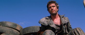 Movie still from “The Road Warrior” (1981), directed by George Miller – A person sitting on the ground near a tire; Close Up shot, Low angle