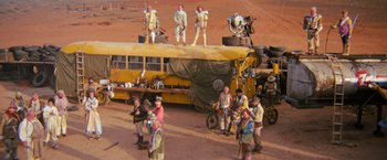 Movie still from “The Road Warrior” (1981), directed by George Miller – A group of people standing next to an old school bus; Extreme Wide shot, High angle