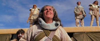 Movie still from “The Road Warrior” (1981), directed by George Miller – An older woman with a scarf around her neck and a man standing on top of a building; Medium shot, Low angle