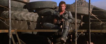 Movie still from “The Road Warrior” (1981), directed by George Miller – A man sitting on top of a pile of tires; Wide shot, Low angle