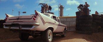 Movie still from “The Road Warrior” (1981), directed by George Miller – An old car with a motorcycle on top of it parked on a dirt field; Wide shot, Low angle