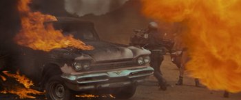 Movie still from “The Road Warrior” (1981), directed by George Miller – An old car on fire with a motorcycle behind it; Wide shot, Low angle