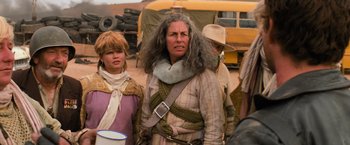 Movie still from “The Road Warrior” (1981), directed by George Miller – A group of people standing next to each other on a dirt field; Medium shot, Over the shoulder angle