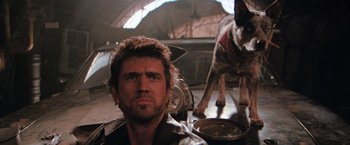 Movie still from “The Road Warrior” (1981), directed by George Miller – A man and a dog are sitting on the floor; Close Up shot, Low angle