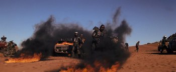 Movie still from “The Road Warrior” (1981), directed by George Miller – A group of men standing next to each other in front of a fire; Wide shot, Low angle