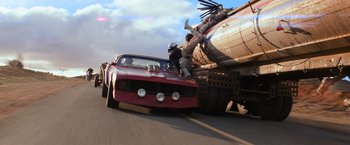 Movie still from “The Road Warrior” (1981), directed by George Miller – A man riding on the back of a red car next to a tanker truck; Wide shot, Low angle