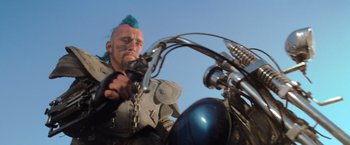 Movie still from “The Road Warrior” (1981), directed by George Miller – A man with a blue mohawk on a motorcycle; Close Up shot, Low angle