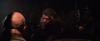 Movie still from “Mad Max Beyond Thunderdome” (1985), directed by George Miller – A man with long hair holding a gun in a dark room; Close Up shot, Over the shoulder angle