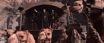 Movie still from “Mad Max Beyond Thunderdome” (1985), directed by George Miller – A man standing on top of a stage next to a camel; Wide shot, Low angle